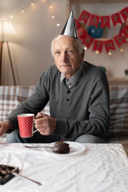 Doğum gününü yalnız kutlayan, parti şapkası takmış, hüzünle kameraya bakan emekli bir adamın portresi. Boşluğu kopyala