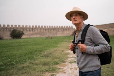 Sırt çantalı yaşlı bir adamın portresi taş duvarların arkasında duran eski bir kalede yaşlı bir turist. Yakın plan.