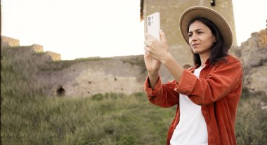 Şapkalı genç bir kadın eski tarihi kalenin yakınında akıllı telefonuyla fotoğraf çekiyor, seyahat anılarını yakalıyor, turistik yerleri geziyor, antik mimari, turizm ve macera konseptini keşfediyor.