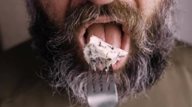 Sakallı adamın Dorblu peyniri yediği ve çataldan bir parça çıkardığı yakın plan. Kırpılmış görünüm