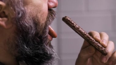 Gri arka planda izole edilmiş çikolata yiyen aç sakallı adam, yakın plan. Yan görünüm