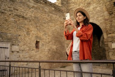 Şapkalı turist kadın cep telefonuyla fotoğraf çekiyor ortaçağ kalesinde taş duvarın karşısında duruyor. Boşluğu kopyala