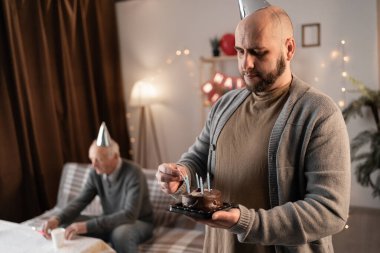 Yaşlı babası evde özel bir dönüm noktasını kutlamak için kanepede otururken sakallı, mutlu, orta yaşlı bir adam pastada mum yakıyor..