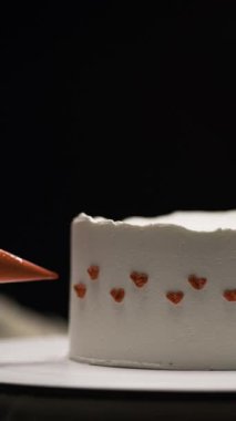 Pasta şefi küçük kalp dekorasyonları yapmak için boru torbası kullanıyor. Beyaz pasta üzerine kırmızı tereyağlı pasta. Karmaşık krema detayı, romantik tatlı, profesyonel pasta dekorasyonu. Dikey video