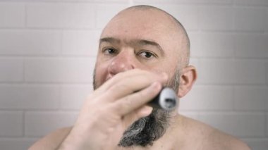 Adam banyoda aynanın yanındaki bıyığı ve sakalı kesmek için elektrikli tıraş makinesi kullanıyor. Yakın plan.