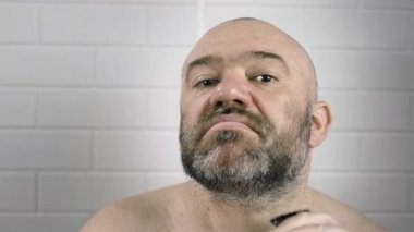 Sakallı adam evde banyoda aynanın önünde makasıyla sakalını kesiyor. Yakın plan.