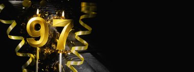 Doksan yedi numaralı altın mum, 97 'nci yaş gününü kutlarken ışıl ışıl yanıyor. Siyah arka planda şenlikli kurdeleler ve konfeti zarif bir kutlama bayrağı oluşturuyor. Boşluğu kopyala.