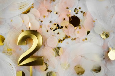 Altın Mum ve Konfeti ile İkinci Doğum Günü Kutlaması. Altın rakamlı iki doğum günü mumu pastel renkli konfetiler, altın yıldız şekilleri ve beyaz tüyler arasında yanıyor..