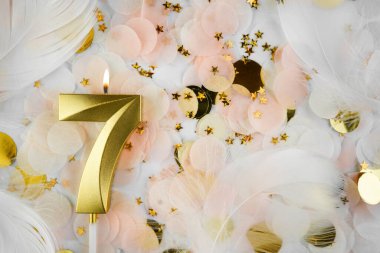 Altın Mum ve Konfeti ile Yedinci Doğum Günü Kutlaması. Pastel renkli konfetiler, altın yıldız şekilleri ve beyaz tüyler arasında altın sarısı bir doğum günü mumu..