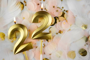 Altın Mum ve Konfeti ile 22. Doğum Günü Kutlaması. Altın rakamlı 22 yaş günü mumu pastel renkli konfeti, altın yıldız şekilleri ve beyaz tüyler arasında yanıyor..