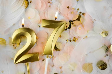 Altın Mum ve Konfeti ile 27. doğum günü kutlamaları. Pastel renkli konfeti, altın yıldız şekilleri ve beyaz tüyler arasında altın rakamlı 27 yaş günü mumu..