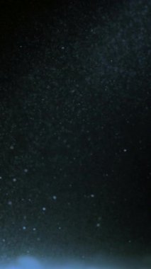 Düşen kar tanelerinin yavaş çekim makrosu yumuşak mavi ışıkla aydınlatılıyor. Hafif kar yağışı rüya gibi bir kış havası yaratıyor. Donmuş atmosfer, dikey 4K video.