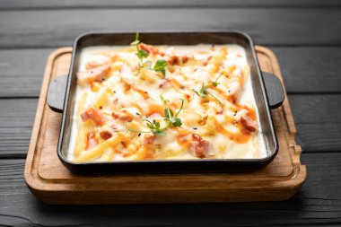 Fırında pişmiş patatesli sıcak dökme demir tava ve kremalı soslu gevrek domuz pastırması, fırından yeni çıkmış, koyu arkaplandan bakıldığında sağlıklı kırsal yemek..