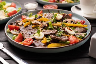 Restoranda ahşap masada servis edilen tabakta ızgara sığır eti, biber, kiraz domatesleri ve bıldırcın yumurtası ile salata.