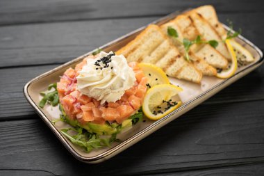 Avokado, krem şanti ve siyah susamlı gurme somon tartar, siyah ahşap masada kızarmış ekmek ve limon dilimleri ile servis edilir.