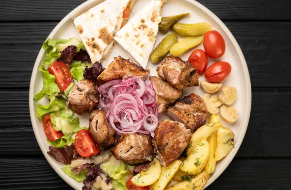 Lavaş, fırında patates, tabakta marine edilmiş soğan ve otlarla servis edilen ızgara etin üst görüntüsü.