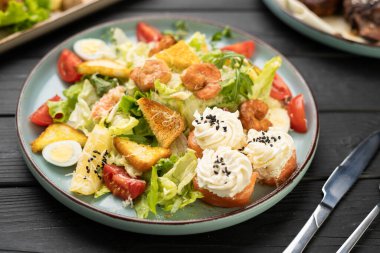 Marul, kiraz, domates, karides, peynir, bıldırcın yumurtası, kızarmış ekmek ve üzerine krem peynirli somon rulosu ve mavi tabakta servis edilen siyah susamlı taze gurme salatası.