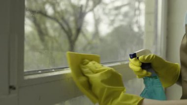 Sarı eldivenli kadın temizlikçi. Pencere çerçevesinden küf çıkarmak için sprey ve bez kullanıyor. Yüzeyi dezenfekte ediyor..