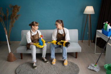 Önlük takan iki kadın hademe ve ellerinde temizlik aletleriyle kanepede dinlenen sarı eldivenler, temizlik sırasında oturma odasında iş tartışmaları, profesyonel temizlik servisi.