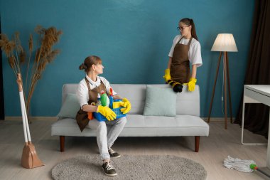 Profesyonel temizlikçi kadın takımı. Kapıcı kanepede oturuyor ve iş arkadaşıyla konuşuyor. Temizlikçiler oturma odasını kanepeyle temizler. Boşluğu kopyala