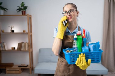 Önlük giymiş temizlikçi kadın sepetin içinde temizlik malzemelerini tutuyor oturma odasında duruyor iş arkadaşıyla telefon görüşmesi yapıyor. temizlik kavramı