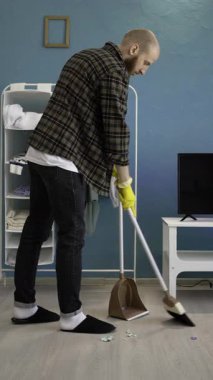 Sakallı adam süpürgeyle temizlik yapıyor ve evdeki oturma odasında yerleri süpürüyor. Temizlik ve temizlik konsepti. Dikey video