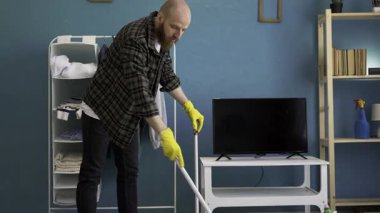 Sakallı adam süpürge ve süpürgeyle evdeki oturma odasında yerleri süpürüyor. Temizlik, ev işi ve temizlik konsepti