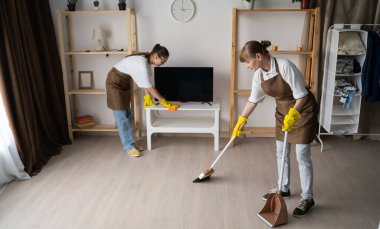 Evi temizleyen, tozu silen ve oturma odasını süpüren iki kadın hademe. Temizlik ve temizlik hizmetleri