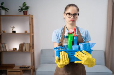 Önlük giymiş, eldiven tutan, temizlik malzemesi taşıyan bir hademe müşterinin evinde durmuş, suratında kötü bir ifade olan kameraya bakıyor. Boşluğu kopyala
