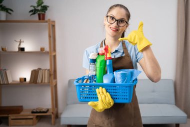 Önlük giymiş, eldiven tutan, temizlik malzemesi taşıyan bir hademe. Müşterinin evinde oturmuş, parmak işareti yapıyor. Boşluğu kopyala