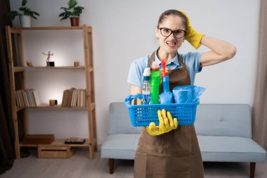 Önlük giymiş, eldiven giymiş, elinde temizlik malzemesi olan üzgün bir kadın hademe. Oturma odasında el ele tutuşup duruyor. Boşluğu kopyala