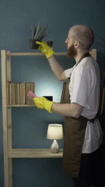 Sakallı adam önlük giyip evi temizliyor. Erkek temizleyici oturma odasında mikrofiber bez kitaplıkla siliyor. Dikey video