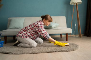 Orta yaşlı ev hanımı, parlak oturma odasında el fırçası kullanarak halı fırçalıyor, ev temizliği, ev işleri, temizlik, temizlik ve günlük temizlik rutinini temsil ediyor.