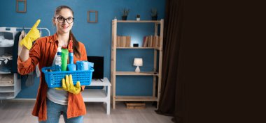 Parlak oturma odasında neşe ve reklam kavramını gösteren temizlik malzemeleriyle dolu bir sepetle gülümseyerek boş bir yeri işaret eden mutlu kahya. Uzayı kopyala Pankart