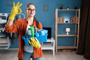 Kadın gülümseyip, parmaklarıyla iyi niyet gösterirken, elinde temizlik ürünleri sepeti tutarak çalışmada olumlu, güven ve memnuniyeti ifade ediyor. Boşluğu kopyala
