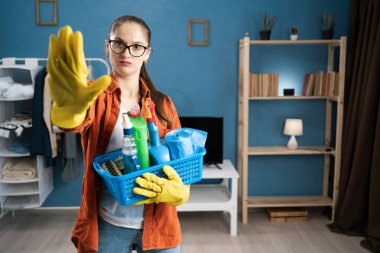 Eli önde olan ciddi bir kadın, elinde temizlik sepetini tutarken, ev içi etkileşim sırasında reddedişini, reddedişini ve sınırlarını gösteren bir jest yapıyor. Boşluğu kopyala