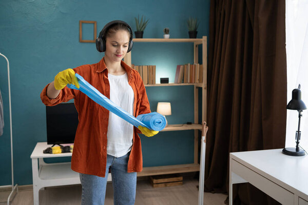 Домработница в наушниках разворачивает рулон мусорных мешков во время выполнения домашней работы и подготовки к домашней уборке