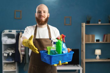 Temizlik malzemeleriyle dolu sepeti tutan sakallı hademe el hareketi yapıyor. Boşluğu kopyala