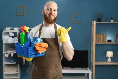 Temizlik malzemeleriyle dolu sepeti tutan sakallı hademe mesaj için parmağını uzayda gösteriyor. Temizlik, temizlik ve ev işi konsepti