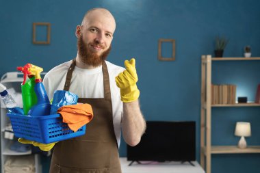 Mutlu Hizmetli sakallı adam sepetle aşk jesti gösteren temizlik malzemelerini tutuyor. Temizlik, temizlik ve ev işi konsepti