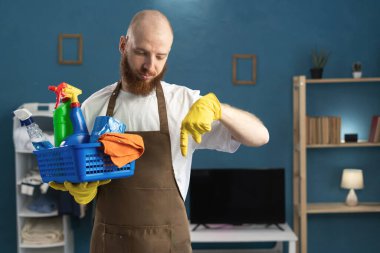 Kapıcı sakallı adam, elinde önlük, elinde temizlik malzemeleri, parmaklarıyla reklamını gösteriyor. Temizlik, temizlik ve ev işi konsepti