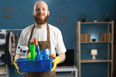 Gülümseyen, sakallı, önlük takan, temizlik malzemesi taşıyan, kameraya bakan mutlu hizmetli. Temizlik, temizlik ve ev işi konsepti