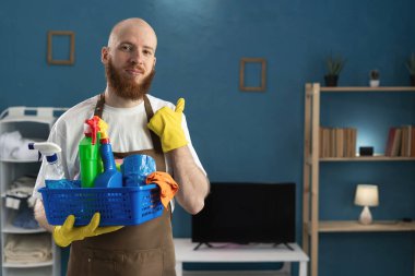Gülümseyen, sakallı, önlük takan, temizlik malzemeleriyle dolu bir sepet taşıyan mutlu hizmetli. Temizlik, temizlik ve ev işi konsepti