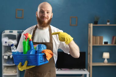 Gülümseyen, sakallı, önlük takan, temizlik malzemesi taşıyan, kendini işaret eden olumlu bir hademe. Temizlik, temizlik ve ev işi konsepti