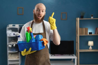 Temizlik malzemeleriyle dolu önlük giyen sakallı hademe, parmak göstererek şaşırdı ve şaşırdı. Temizlik, temizlik ve ev işi konsepti
