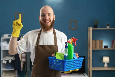 Neşeli, sakallı, önlük giyen, temizlik malzemesi taşıyan, fikir jesti yapan, zaferi kutlayan neşeli hademe sakallı adam. ev işi ve temizlik konsepti