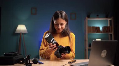 Genç esmer kadın evde oturuyor ve fotoğraf makinesine harici flaş takıyor. Fotoğraf düzeneği, teknik hazırlık, yaratıcı hobi ve modern fotoğrafçının çalışma alanı.