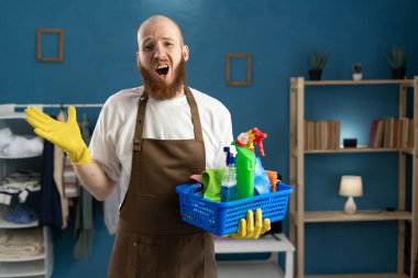 Yakışıklı sakallı adam elinde süpriz bir ifadeyle temizlik ürünleri tutuyor özür diliyor, oturma odasında dikilirken özür diliyor. Ev işleri, ev işleri, ev işleri ve temizlik konsepti.