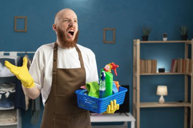 Yakışıklı, sakallı, kel bir adam temizlik malzemelerini tutuyor, oturma odasında dururken kollarını açıp öfkeyle bağırıyor. Ev işleri, ev işleri, ev işleri ve temizlik konsepti..