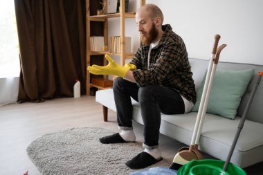 Adam oturma odasında kanepede otururken temizlik için sarı lastik eldiven giyiyor. Ev işleri. Boşluğu kopyala
